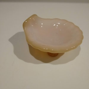 May include: A small, white, glass dish shaped like a seashell with a scalloped edge. The dish has a slightly iridescent finish and sits on a small, round, brown base.