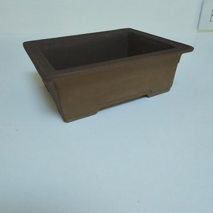 May include: A rectangular brown ceramic bonsai pot with a slightly raised rim and four small feet.