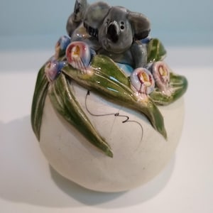 May include: A ceramic figurine of a koala sitting on a bed of green leaves and pink flowers. The koala is gray and has a white belly. The figurine is hand-painted and has a white glaze.