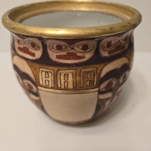 May include: A small, round ceramic pot with a gold rim. The pot features a repeating pattern of stylized faces and geometric shapes in shades of brown, beige, and red. The interior of the pot is a light, translucent color.