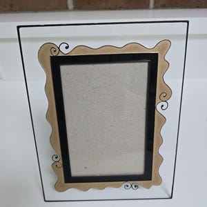 May include: A rectangular picture frame with a black border and a decorative gold-colored inner frame. The frame has a clear glass front and a black wire frame. The gold frame has decorative swirls.