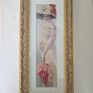 May include: A framed vintage print of a woman in a hat and dress, holding a pink parasol. The print is in a gold ornate frame. The woman is wearing a large hat with red accents and a light-colored dress.