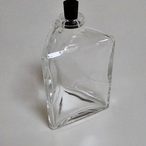 May include: A clear glass bottle with a black plastic stopper. The bottle has a rectangular shape with rounded corners.