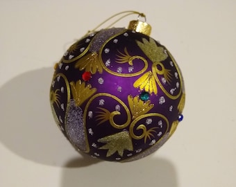 May include: Purple Christmas ornament with gold swirls and glitter accents. The ornament has small, colorful jewels embedded in the design.