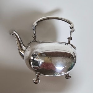 May include: A silver teapot with a hinged handle and three feet. The teapot is polished and reflects the surrounding environment.