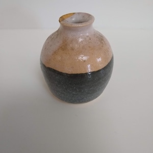 May include: A small, round ceramic vase with a light brown and black glaze. The vase has a wide mouth and a narrow neck.
