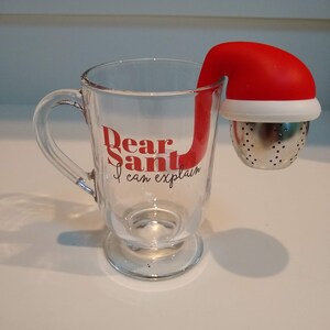 May include: A clear glass mug with a handle, featuring the text "Dear Santa I can explain". A red and white Santa hat shaped tea infuser is attached to the mug.