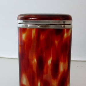 May include: A rectangular, vintage case with a tortoiseshell pattern in shades of red, brown, and cream. The case has a silver-colored metal trim around the top and a hinged lid. The overall design is classic and elegant.