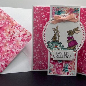 Pop up Klappkarte Häschen mit Bilderbuch. Rosa und pastellrosa Blüten. Stimmungs- (Vorderseite) Ostergrüße, (innen)Ein Häschen Liebt Dich.