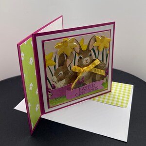 Saudações de Páscoa. Dois coelhos em narcisos. Fundo magenta, branco e flores. Interior - em branco com pequeno narciso pequeno.
