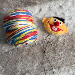 May include: A colorful ball of yarn with red, yellow, blue, and white stripes sits next to a plush, smiling, yellow character head with a red nose, black hair, and a pink mouth. A black string is attached to the character's head.