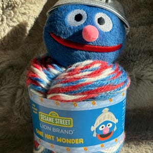 May include: A blue Grover soft toy wearing a silver helmet, on top of a spool of yarn. The yarn is red, white, and blue. The spool has the text "Sesame Street" and "One Hat Wonder".