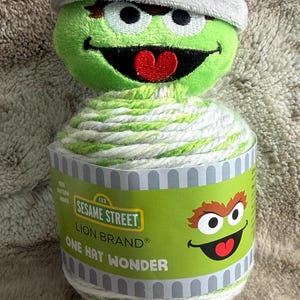 May include: A plush Oscar the Grouch head sits atop a skein of Lion Brand yarn. The yarn is white and green. The label reads "Sesame Street One Hat Wonder". The plush Oscar has a gray hat and a red tongue.