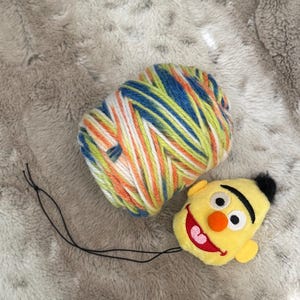 May include: A skein of colorful yarn in blue, green, orange, and white sits next to a yellow plush toy shaped like a cartoon character's head. The toy has a black hair tuft, orange nose, and a red smiling mouth. A black string is attached to the toy.