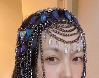 May include: A woman wearing a silver chain headpiece with iridescent blue and clear crystals. The headpiece is made of multiple chains and hangs down her back. She is wearing a light green sleeveless shirt.