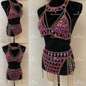 May include: A gold chain body jewelry set with pink iridescent rhinestones. The set includes a bra top and a skirt with fringe.