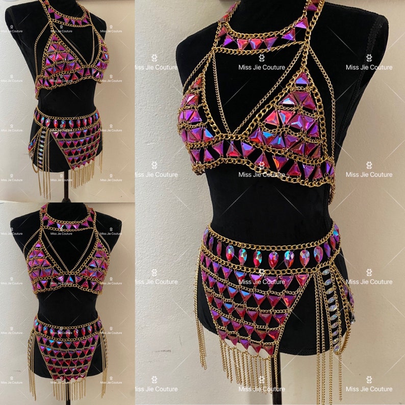 May include: A gold chain body jewelry set with pink iridescent rhinestones. The set includes a bra top and a skirt with fringe.