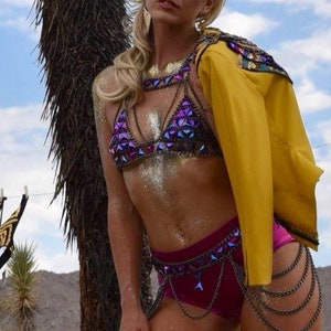 May include: A woman wearing a purple and silver sequined bikini top with a yellow jacket and a silver chain belt. The woman is standing in front of a palm tree.
