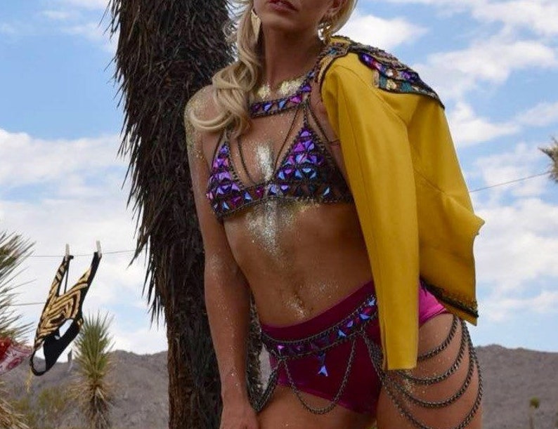 May include: A woman wearing a purple and silver sequined bikini top with a yellow jacket and a silver chain belt. The woman is standing in front of a palm tree.
