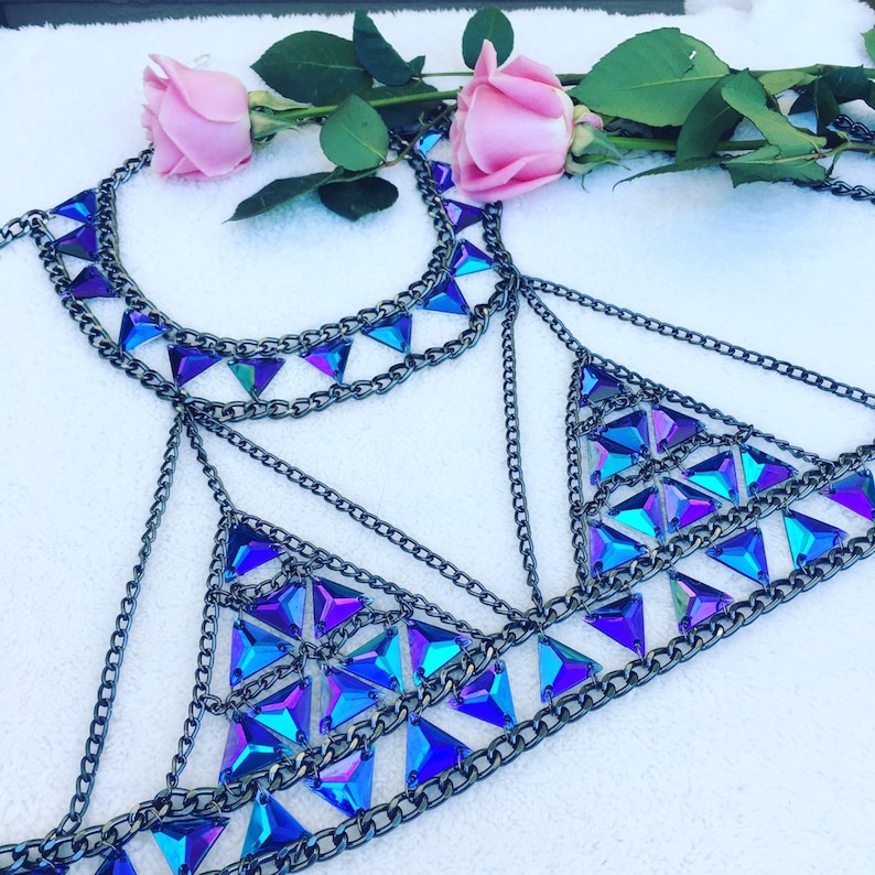 May include: A silver chain body jewelry piece with iridescent blue and purple triangle-shaped rhinestones. The jewelry is on a white surface with pink roses.