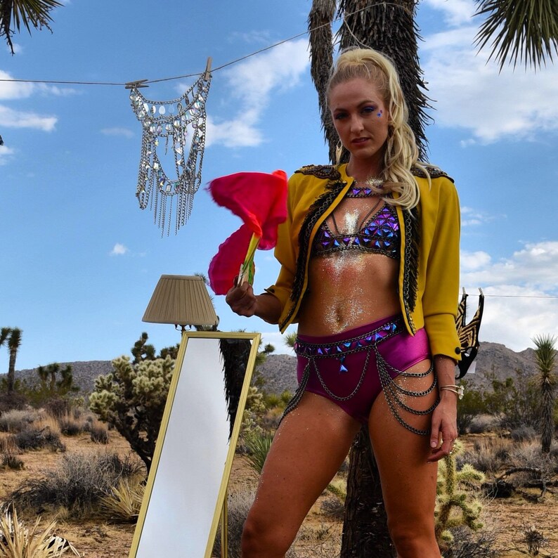May include: A woman wearing a yellow jacket, a purple and blue sequined bra, and a purple skirt with a chain belt. She is holding a pink feather boa and standing in front of a mirror.
