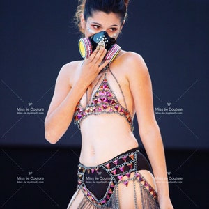 May include: A woman wearing a black and pink chainmail bikini top and skirt with a black gas mask with pink and yellow accents.