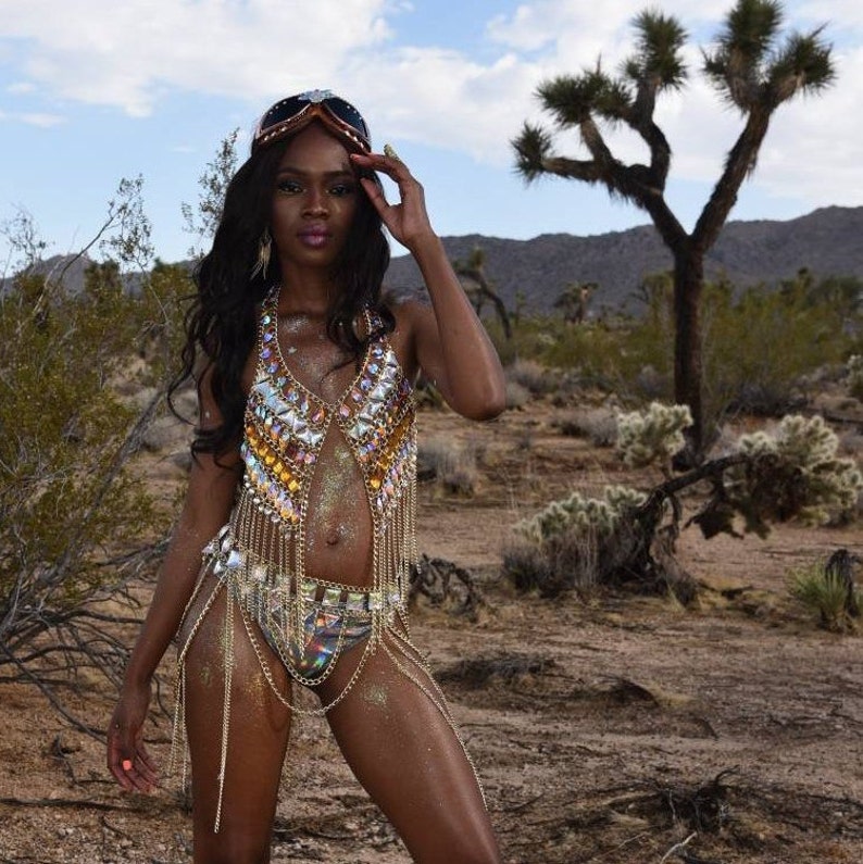 May include: A woman wearing a gold and silver chainmail top with iridescent rhinestones and a matching skirt. She is wearing a pair of goggles and is standing in a desert landscape.