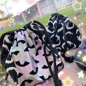 May include: Two black fabric drawstring bags with different patterns. One bag has a white and black pattern of fangs and the other has a silver and black pattern of bats.
