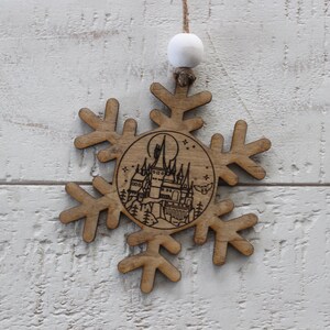 May include: A wooden snowflake ornament with a laser-engraved image of a castle and a crescent moon. The ornament is brown and has a white string for hanging.