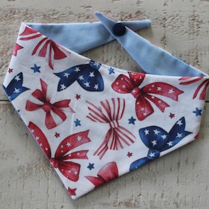 May include: A dog bandana with a patriotic theme. The white fabric is patterned with red and blue bows and stars. The reverse side and the neck ties are light blue. The bandana is secured with a dark blue button.