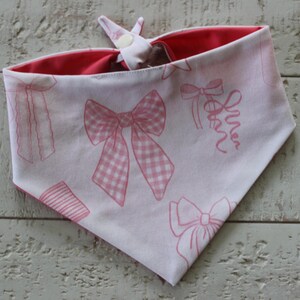 May include: A white and pink dog bandana with a bow pattern. The bandana is reversible, with a solid pink interior. The bow pattern includes gingham and ribbon designs. The bandana is tied at the top with a white fabric and a button.