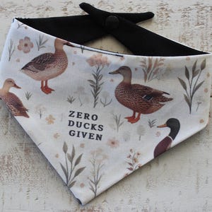 May include: A white dog bandana with a black backing and snap closure. The bandana features a pattern of ducks, flowers, and foliage, with the phrase "ZERO DUCKS GIVEN" in black text.