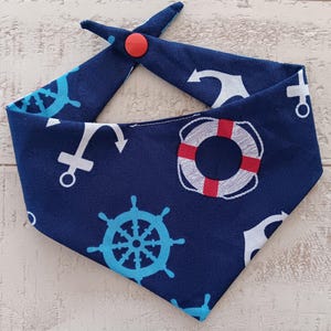 May include: A blue bandana with a nautical theme. It features anchors, life preservers, and steering wheels in white, red, and blue. The bandana has a red button closure.