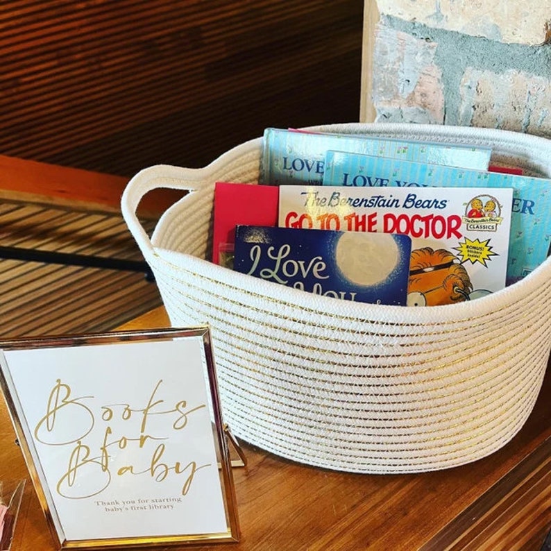 Puede incluir: Una cesta tejida blanca con detalles dorados llena de libros para ni&ntilde;os. Los libros tienen portadas coloridas con t&iacute;tulos como "The Berenstain Bears Go To The Doctor" e "I Love You". Un cartel frente a la cesta dice "Books for Baby. Thank you for starting baby's first library."