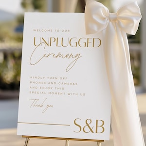 Puede incluir: Un letrero blanco con texto dorado que dice "WELCOME TO OUR UNPLUGGED Ceremony". Solicita a los invitados que apaguen los teléfonos y las cámaras. Un gran lazo color crema adorna la esquina superior derecha. El letrero está sobre un caballete dorado.