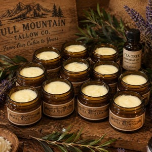 May include: A collection of amber glass jars filled with off-white tallow balm, each featuring a Bull Mountain Tallow Co. label. The jars are arranged with a bottle of tea tree essential oil, sprigs of lavender, and a wooden sign with mountain graphics.