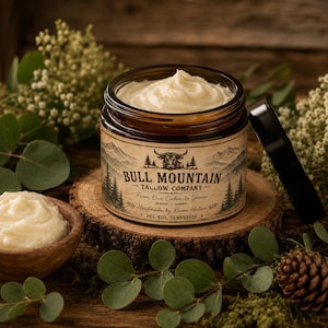 May include: A glass jar filled with white tallow cream, featuring a label that reads Bull Mountain Tallow Company, From Our Cabin to Yours, Handmade by Karen Halion, Del Rio, Tennessee, with a bull illustration and mountain scenery.