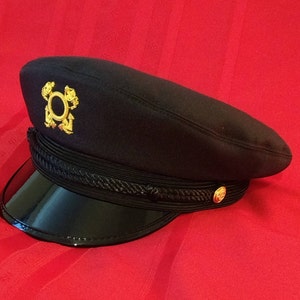 May include: A navy blue hat with a shiny black visor. The hat features a gold emblem and a decorative rope band. A gold button is on the side. The hat is likely part of a uniform or formal attire.