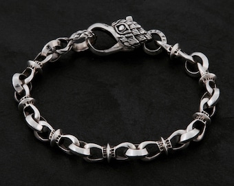 Könnte beinhalten: Ein silbernes Kettenarmband bestehend aus einer Reihe ineinandergreifender runder Glieder mit strukturierten Akzenten. Das Armband wird durch einen Verschluss in Form eines stilisierten Adlerkopfes mit detaillierter Federgravur geschlossen.