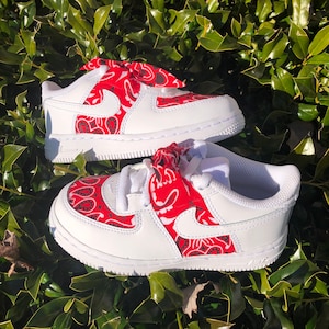 May include: White toddler sneakers with red paisley accents on the sides and tongue. The sneakers feature a white Nike swoosh and white laces. The red paisley pattern has black and white details.