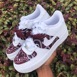 May include: White sneakers with burgundy paisley accents on the toe and side panels. The sneakers feature a white sole, white laces, and a burgundy bow tied at the laces. The Nike swoosh is visible on the side.