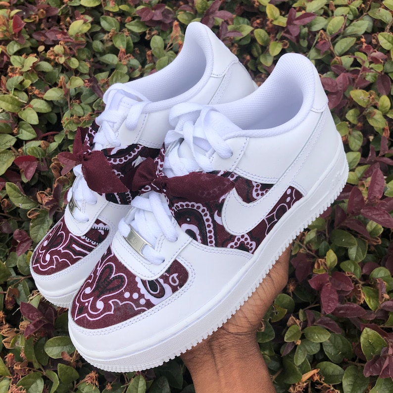 May include: White sneakers with burgundy paisley accents on the toe and side panels. The sneakers feature a white sole, white laces, and a burgundy bow tied at the laces. The Nike swoosh is visible on the side.