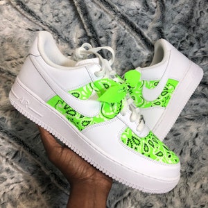 May include: White leather sneakers with neon green paisley pattern accents. The sneakers feature a white sole, laces, and the Nike swoosh logo. A neon green bow is tied to the laces.