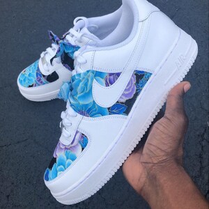 May include: White Nike Air Force 1 sneakers with a floral design in shades of blue and purple. The design is on the sides and toe box of the shoes. The Nike swoosh is white. The shoes are held by a hand.