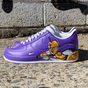 May include: Purple and gold custom Nike Air Force 1 sneakers with a painted image of a Spartan warrior on the side.