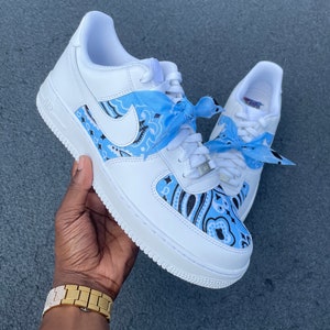 May include: White sneakers with light blue paisley pattern accents and matching light blue laces. The shoes feature a white sole and a white Nike swoosh. The laces are tied with light blue bandanas.