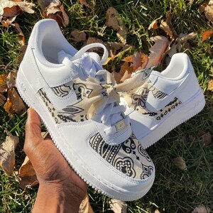 May include: White sneakers with black and tan paisley pattern accents on the sides and tongue. The sneakers have white laces and a tan paisley ribbon tied in a bow. The shoes are on a grassy surface.