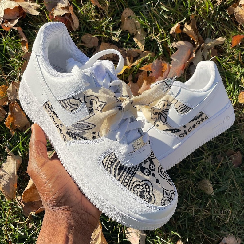 May include: White sneakers with black and tan paisley pattern accents on the sides and tongue. The sneakers have white laces and a tan paisley ribbon tied in a bow. The shoes are on a grassy surface.