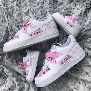 May include: White sneakers with pink paisley design accents and pink bows. The sneakers have the word "AIR" on the side and the names "Dior Kongss" and "Kyri" in pink script. The shoes are on a gray, textured surface.