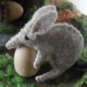 May include: A grey felt toy mouse holding a brown egg. The mouse has a long snout and large ears.